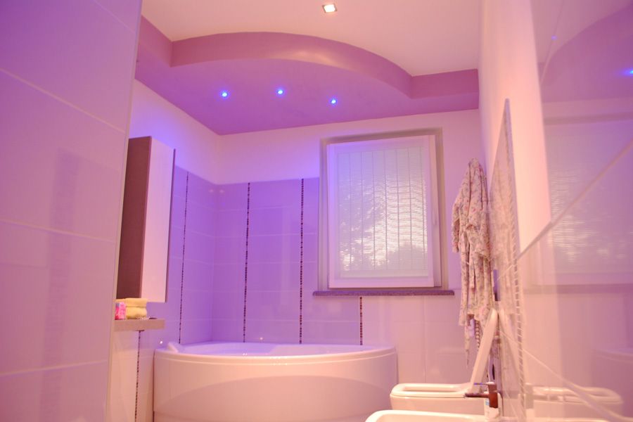 Bagno illuminato di viola con vasca bianca, pareti piastrellate e un'ampia finestra con tende.