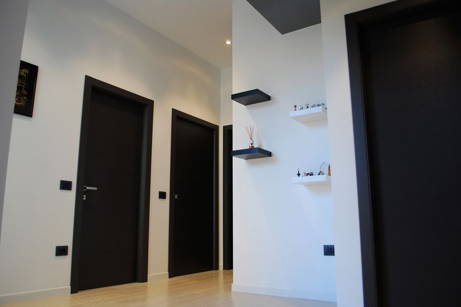 Corridoio moderno con tre porte nere, pavimento in legno chiaro e mensole a muro su una parete bianca.