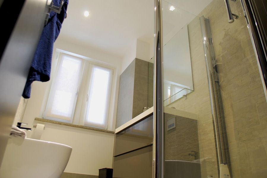 Piccolo bagno con doccia, lavandino, WC e finestra; un asciugamano blu è appeso a sinistra.