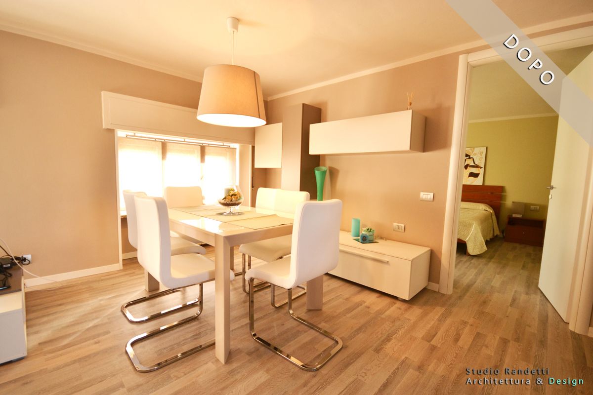 Luminosa zona pranzo con tavolo e sedie bianchi, lampada a sospensione, pavimento in legno e una camera da letto visibile attraverso una porta aperta.