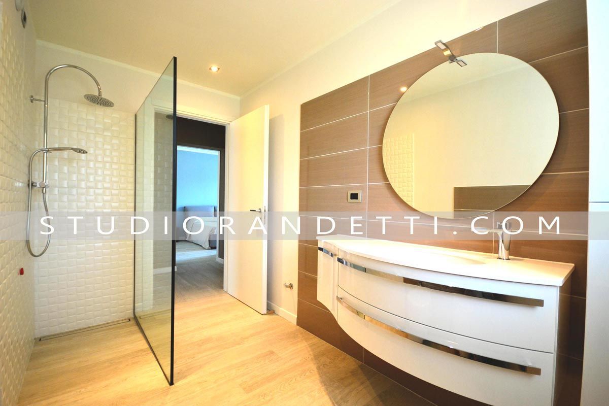 Bagno moderno con cabina doccia in vetro, specchio rotondo e mobile lavabo curvo bianco in calde tonalità di legno.