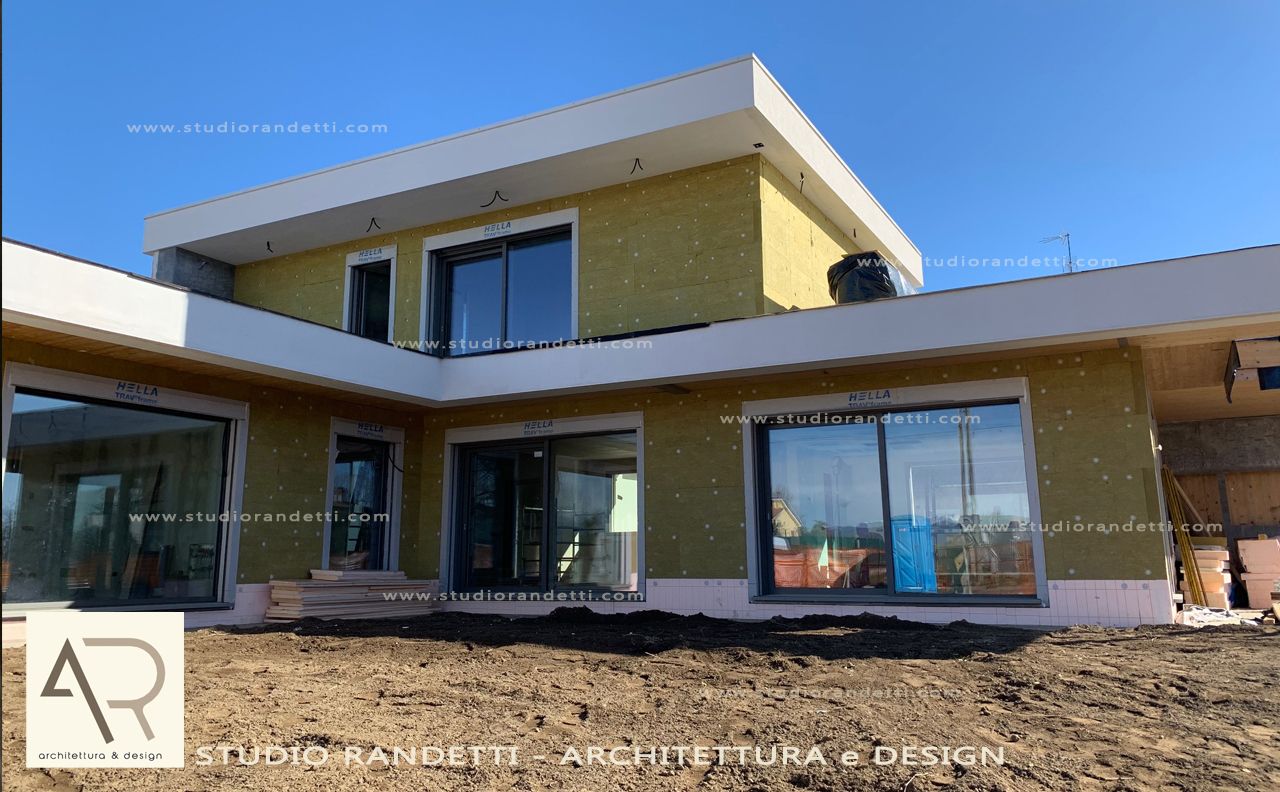 Esterno di una moderna casa a due piani con ampie finestre, intonaco color sabbia e tetto piano sotto un cielo azzurro limpido.
