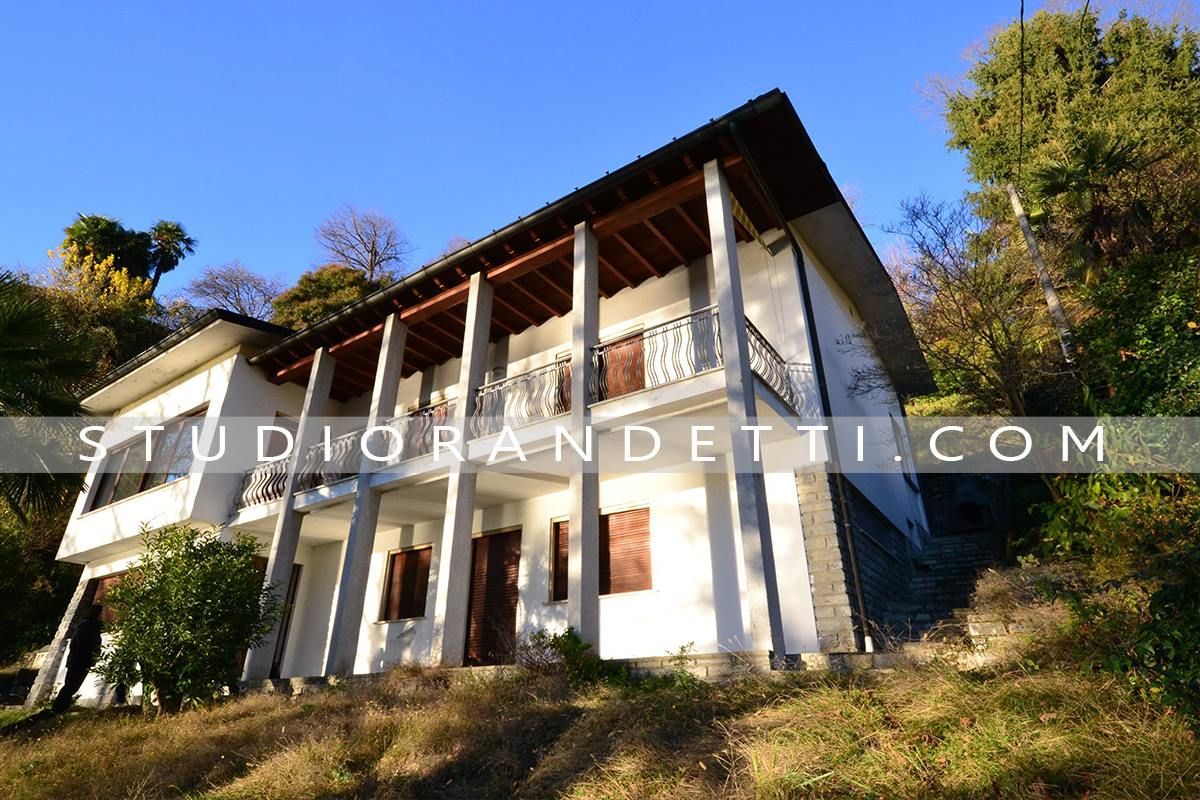 Moderno rifugio di montagna con pareti bianche, balcone in legno e base in pietra su un pendio erboso sotto un cielo azzurro.