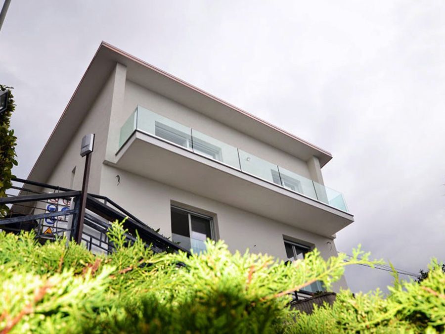 Esterno di una casa moderna con balcone in vetro, visto da un'angolazione bassa, incorniciato dal verde e da un cielo nuvoloso.