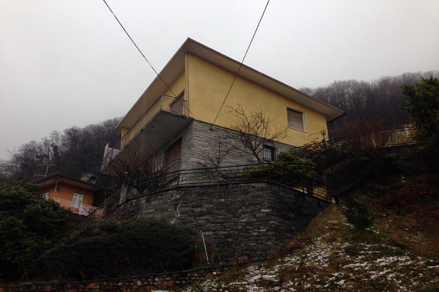 Casa gialla su un ripido pendio con fondamenta in pietra, cielo nuvoloso e alberi spogli.