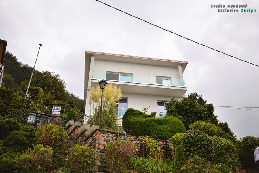 Casa bianca a due piani su una collina con balcone, circondata da una fitta vegetazione sotto un cielo nuvoloso.
