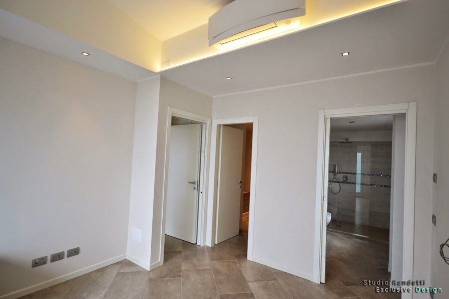 Luminosa stanza moderna e vuota con pareti bianche, faretti a incasso sul soffitto e porte aperte che conducono al bagno e al corridoio.
