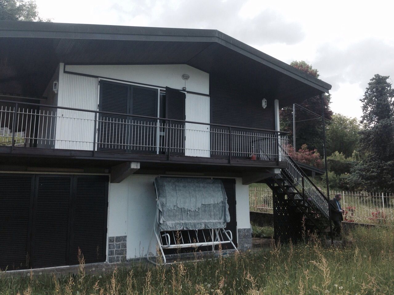 Casa a due piani con balcone, persiane scure e giardino antistante.