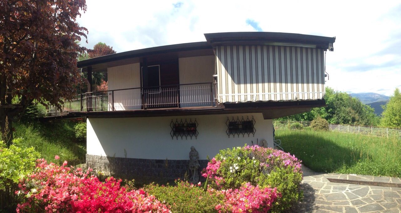 Casa a due piani con balcone e rivestimento esterno bianco, circondata da fiori e vegetazione in un giardino soleggiato.