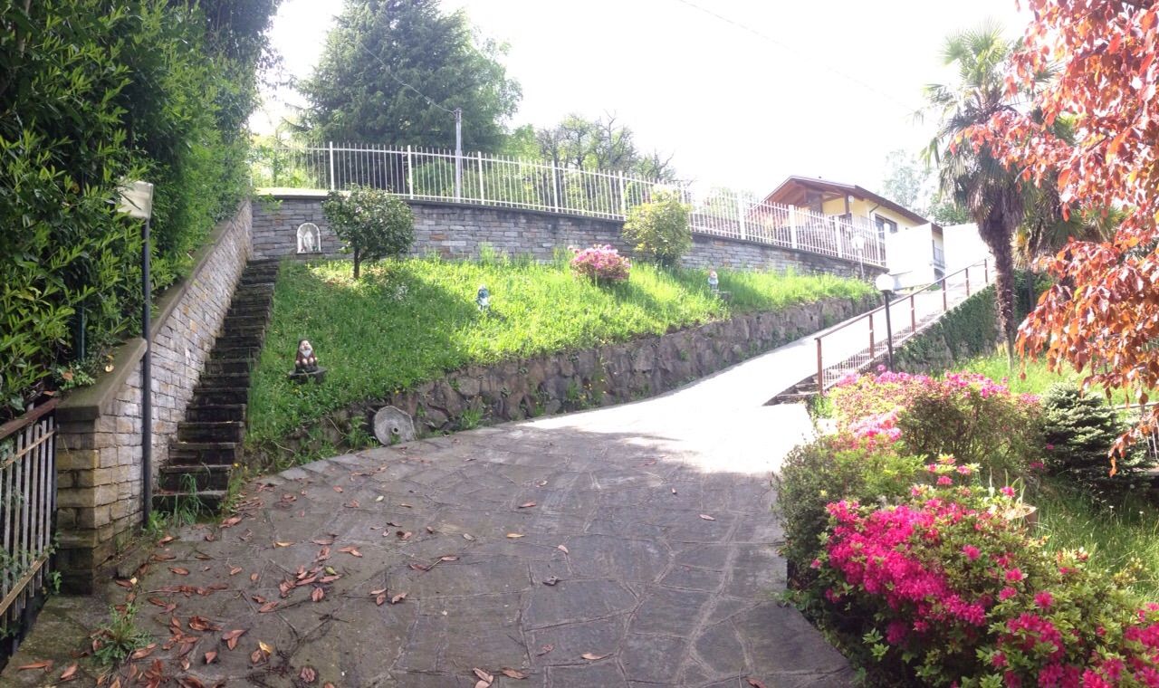 Sentiero curvo in pietra che attraversa un giardino collinare verdeggiante con scalini, fiori rosa e una casa sullo sfondo.