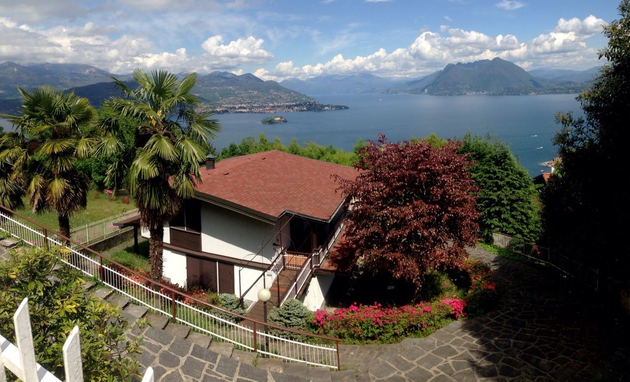 Casa sul lago con tetto rosso, palme e vista su un lago di montagna sotto un cielo parzialmente nuvoloso.