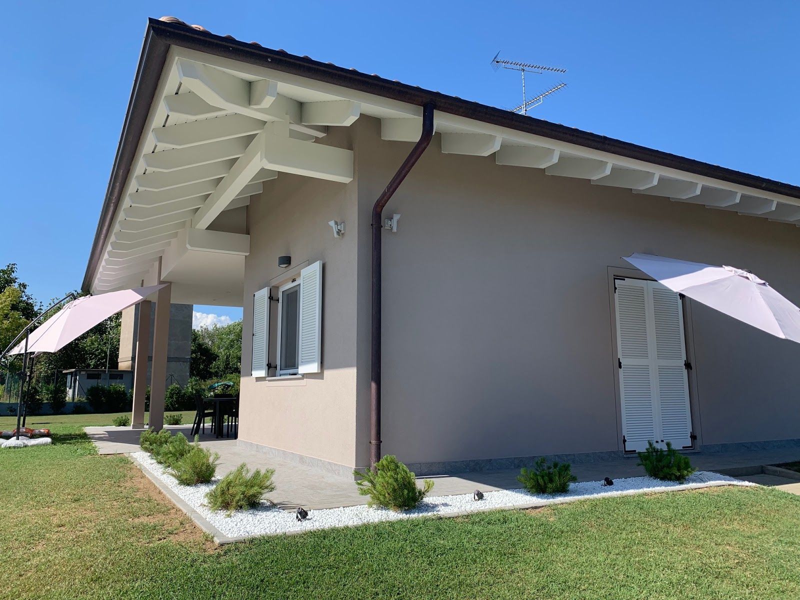 Casa moderna color beige con persiane bianche, tetto spiovente e ombrelloni da giardino su un prato soleggiato.