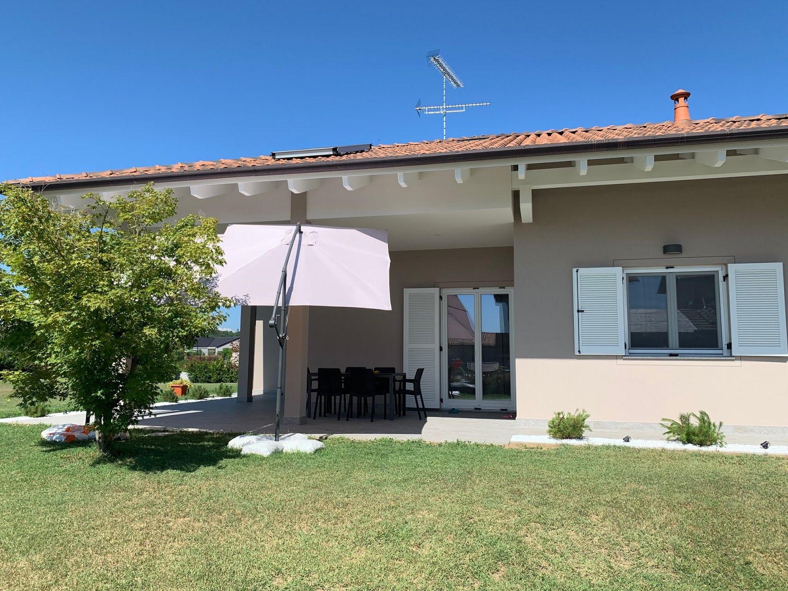 Casa a un solo piano con patio, ombrellone, porta aperta e prato sotto un cielo azzurro.