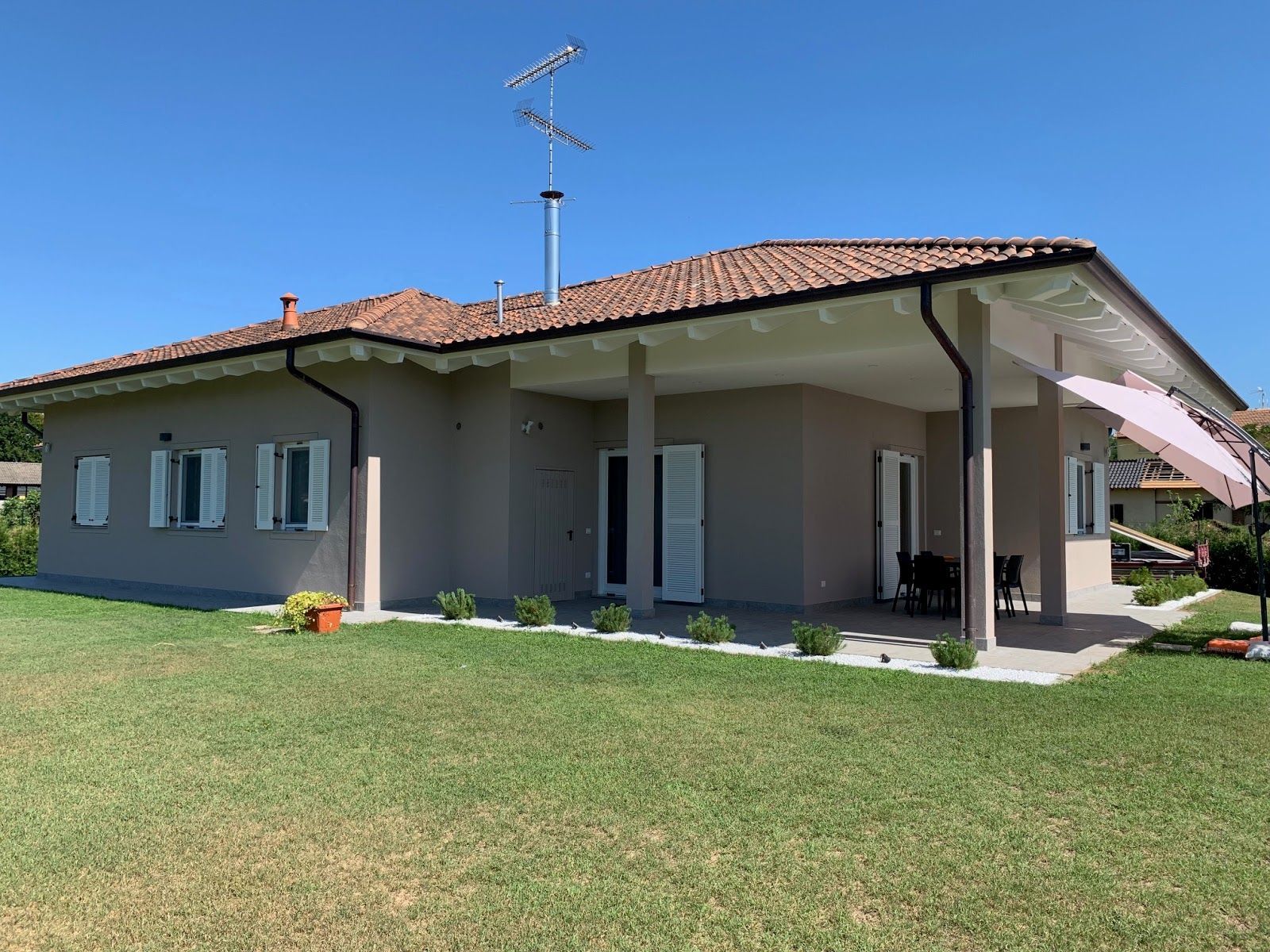 Casa grigia a un solo piano con tetto di tegole rosse, patio coperto e prato verde sotto un cielo limpido