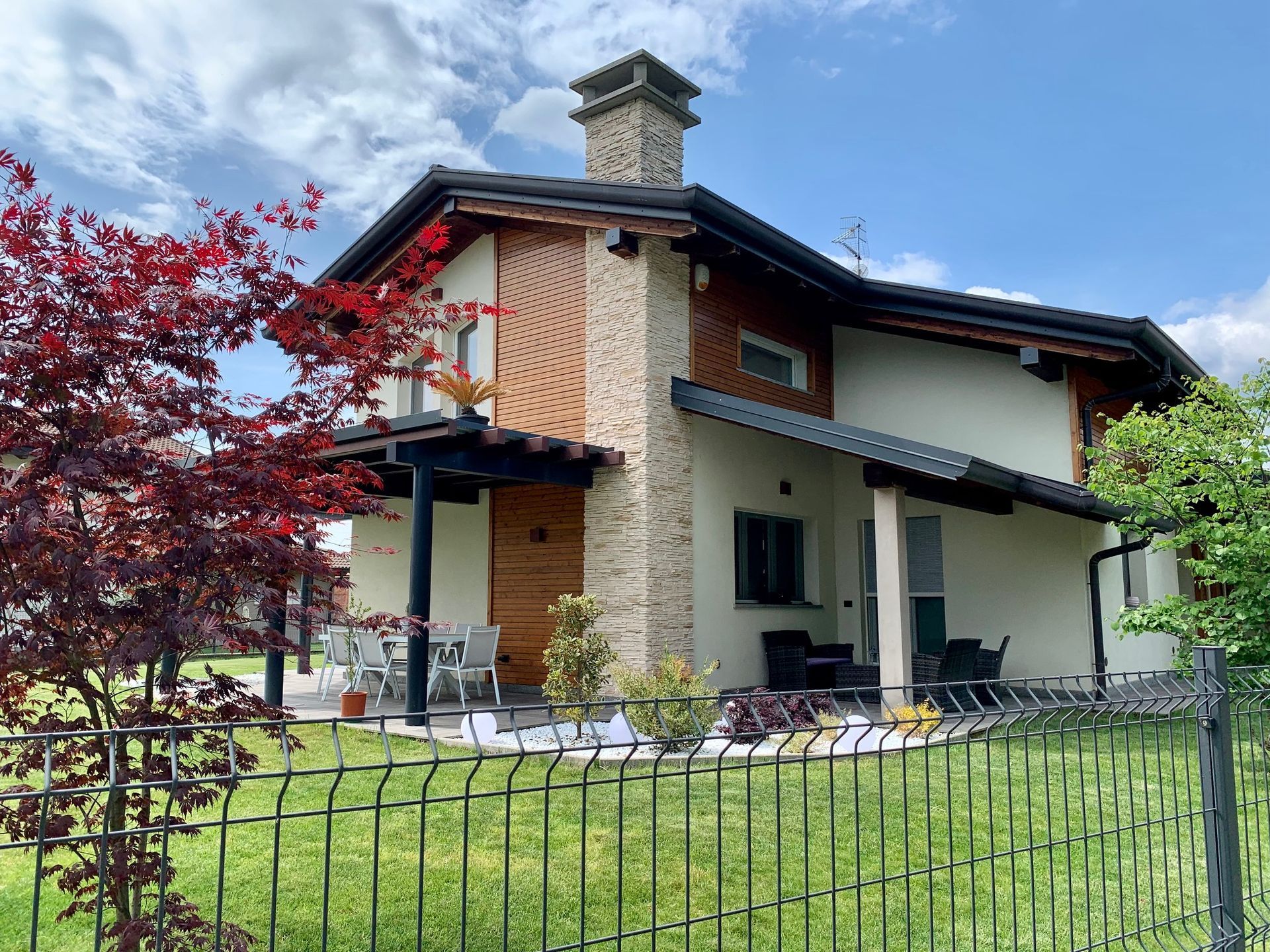 Casa moderna con camino in pietra, albero rosso e recinzione nera in un cortile verde