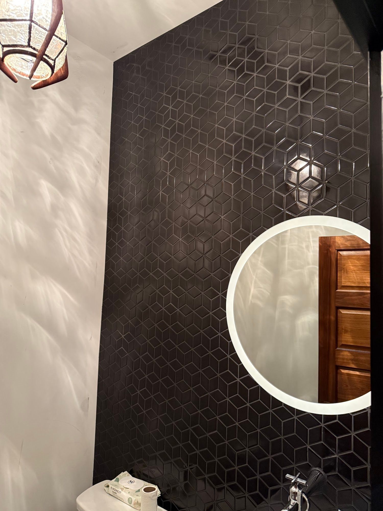 Dark tiled accent wall in a bathroom, with a circular mirror and overhead lamp.