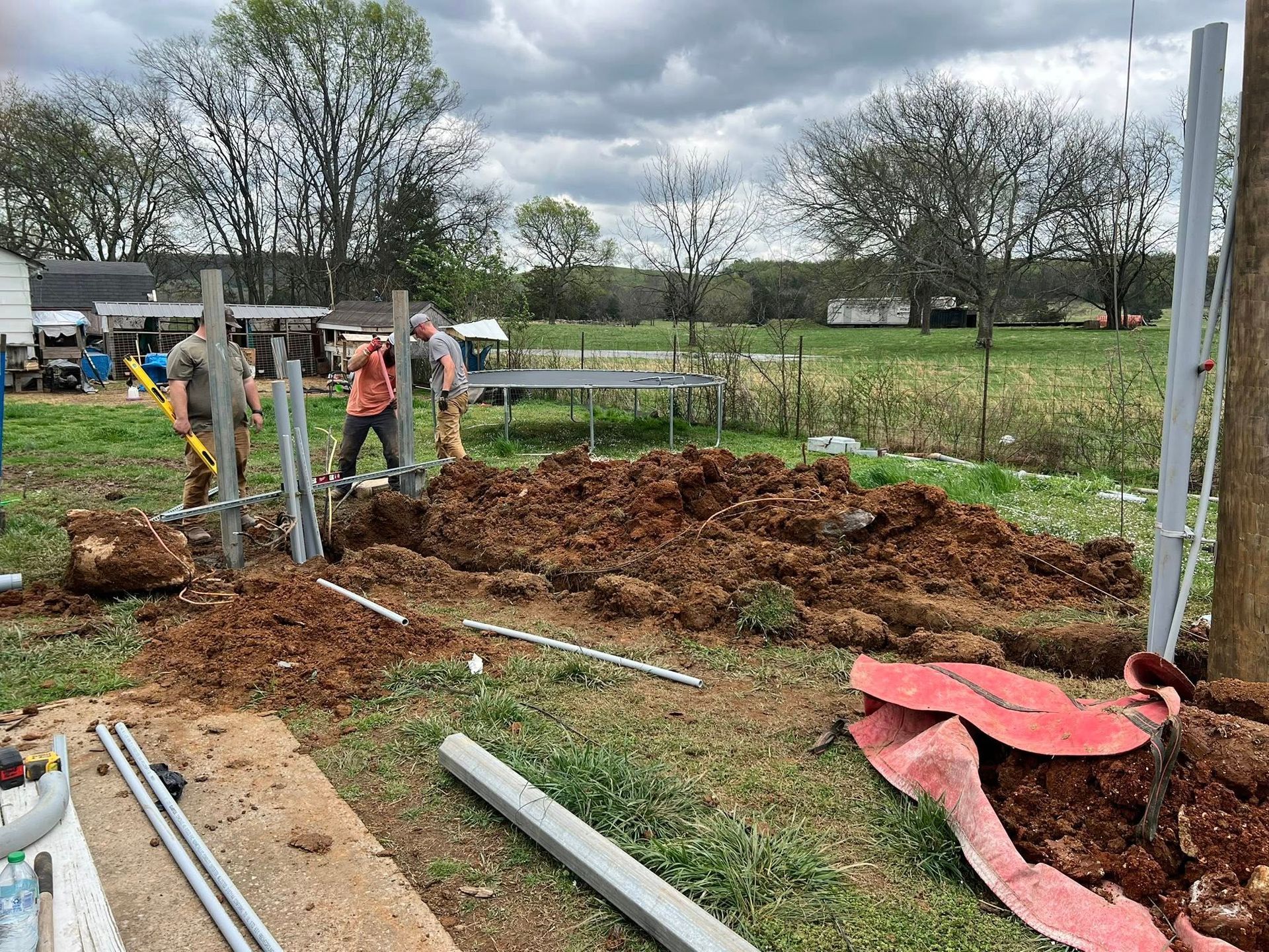 People constructing a structure in a grassy yard. Dirt pile and materials surround workers. Cloudy sky.
