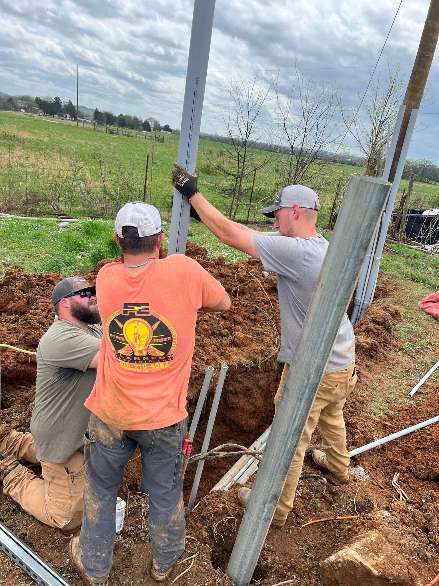 Three workers installing a metal post in a muddy hole in a field. Cloudy sky.