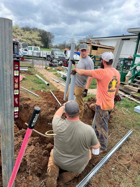Three workers installing a fence, one in a trench holding a post, two others assisting; outdoors.