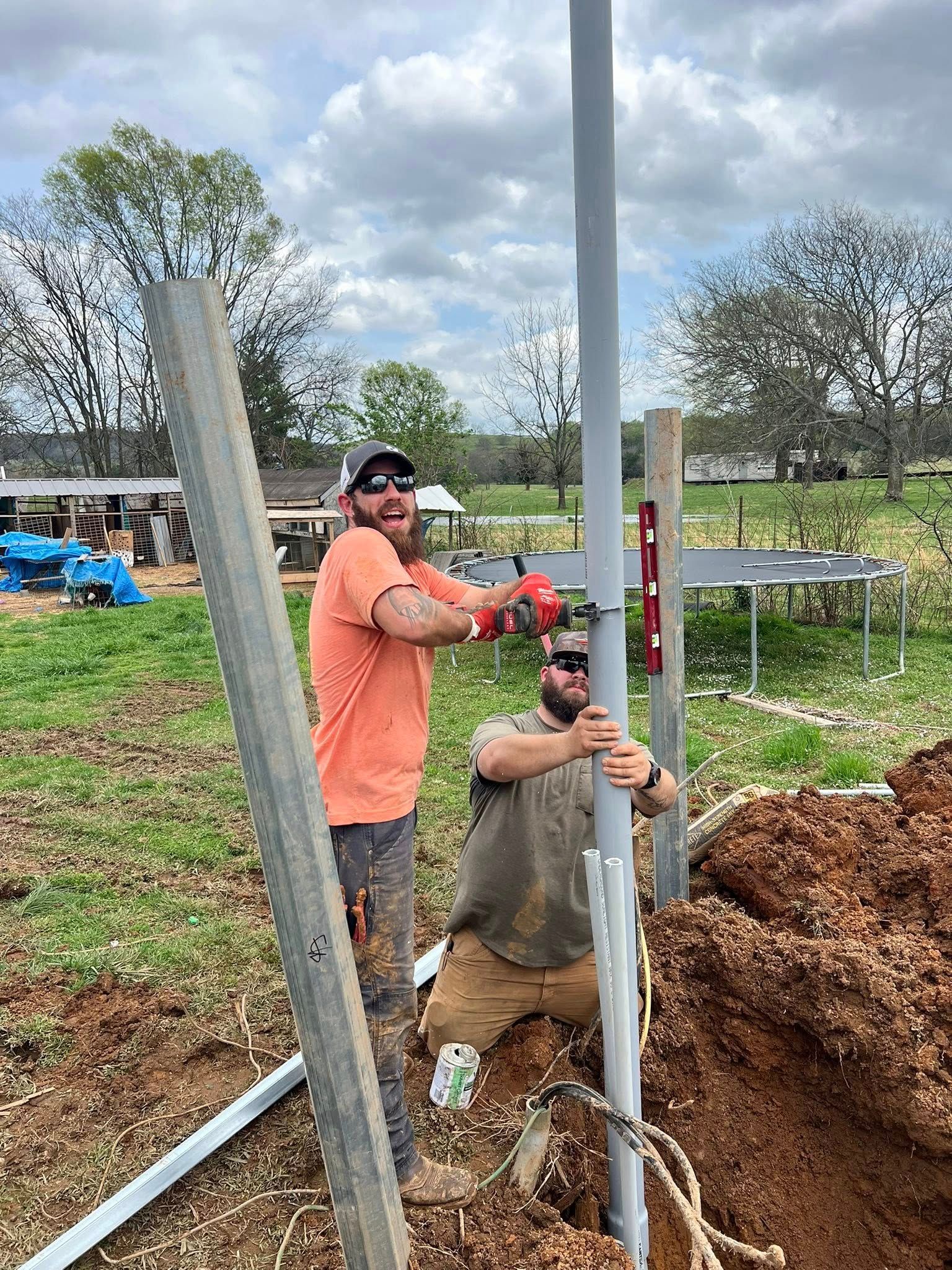 Two men installing a metal pole outdoors, one using a drill, the other kneeling and holding the pole, overcast day.