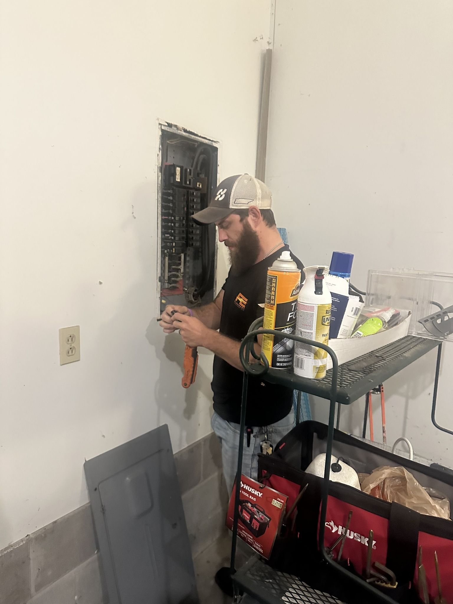 Electrician working on electrical panel, inside a garage. He’s wearing a hat, shorts, and using tools. A toolbox is nearby.