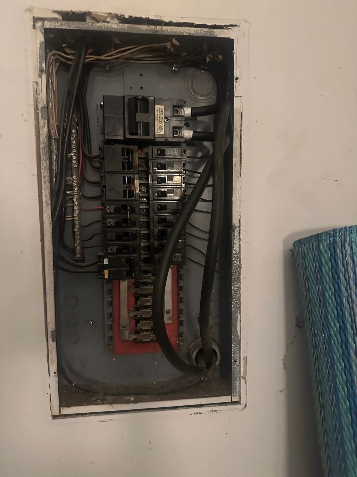 Open electrical breaker panel with exposed wiring in a wall recess, next to a blue rolled mat.