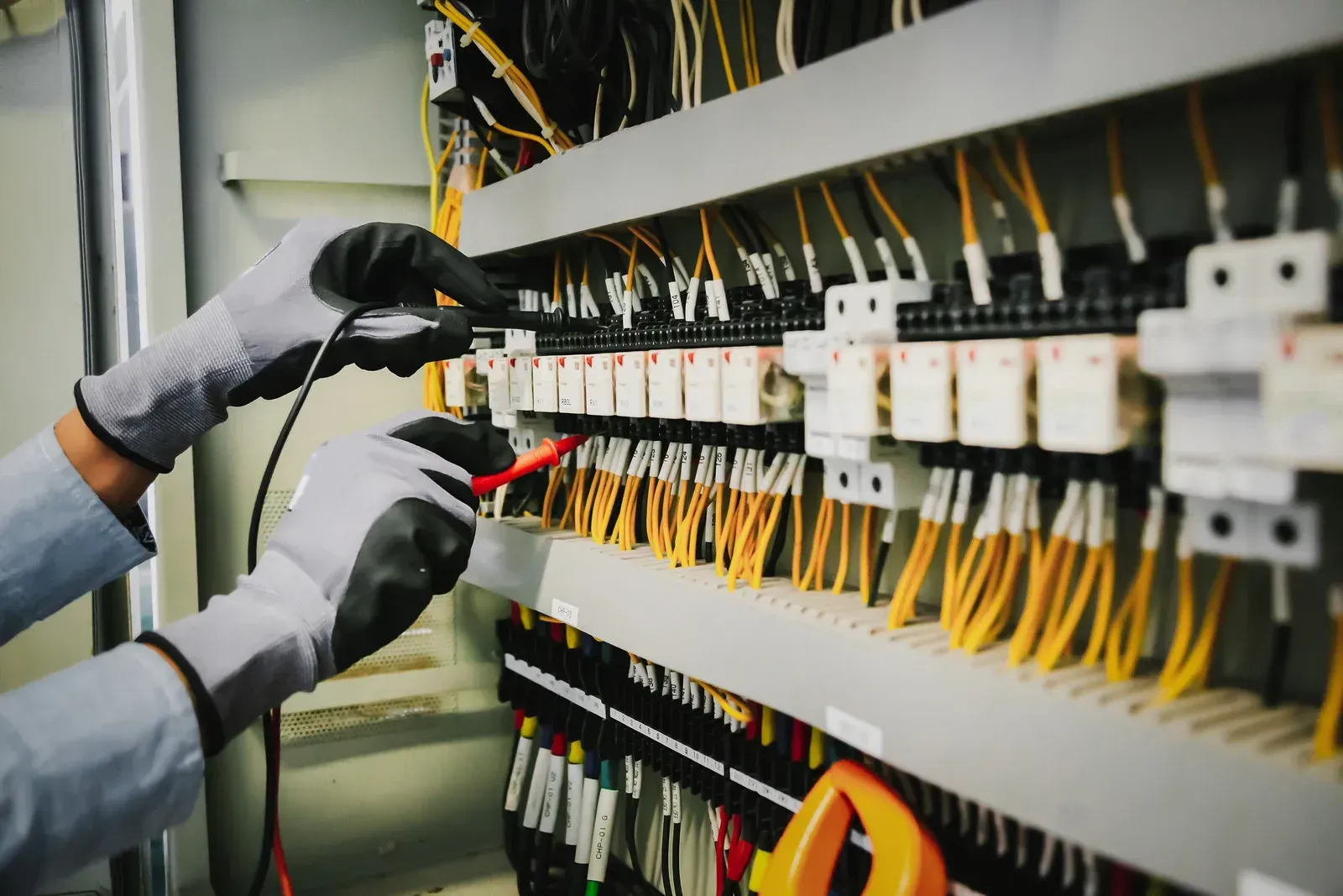 An electrician in safety gloves uses a multimeter to test electrical components inside a control panel with yellow wires.