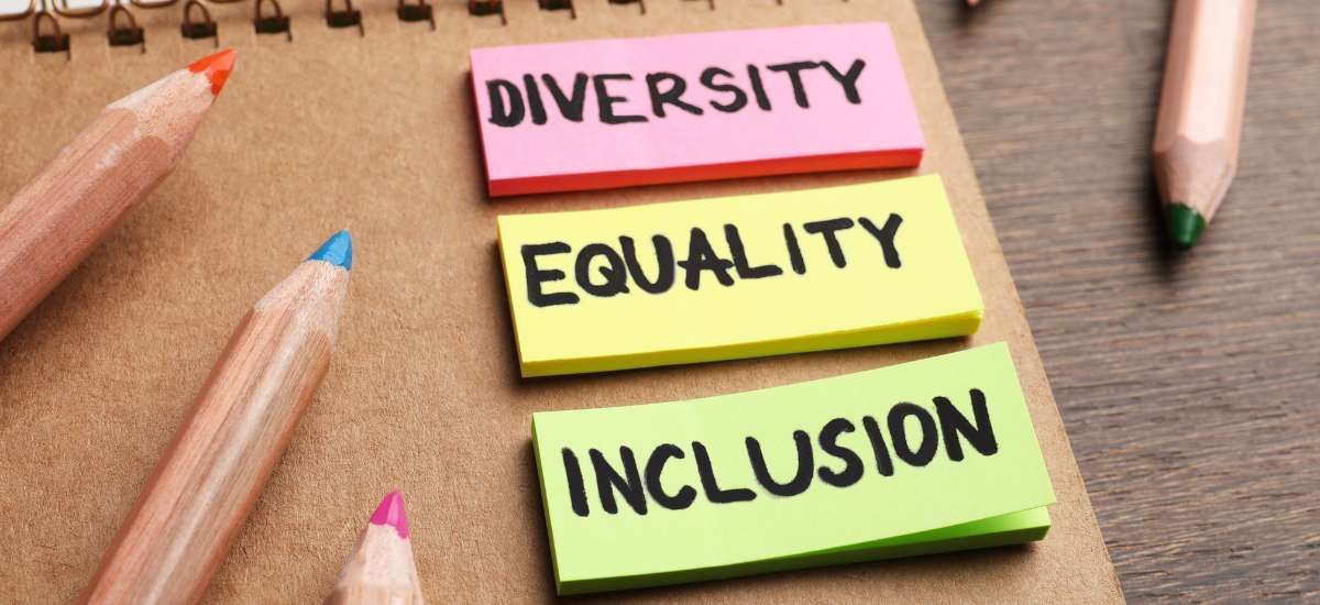 On a desk is a notepad, colored pencil crayons, and 3 different colored sticky notes; each with a single word on it. 1- Diversity, 2- Equality, 3 - Inclusion
