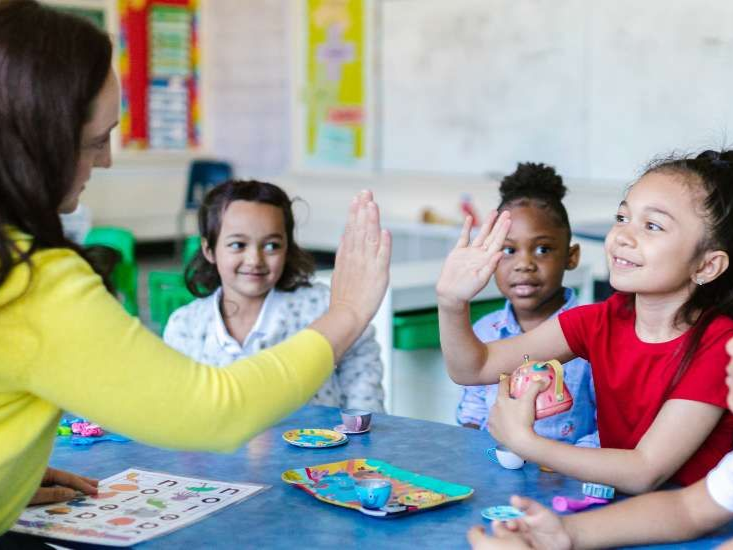 Teacher and 3 students at an art table. She is giving one little girl a high five.