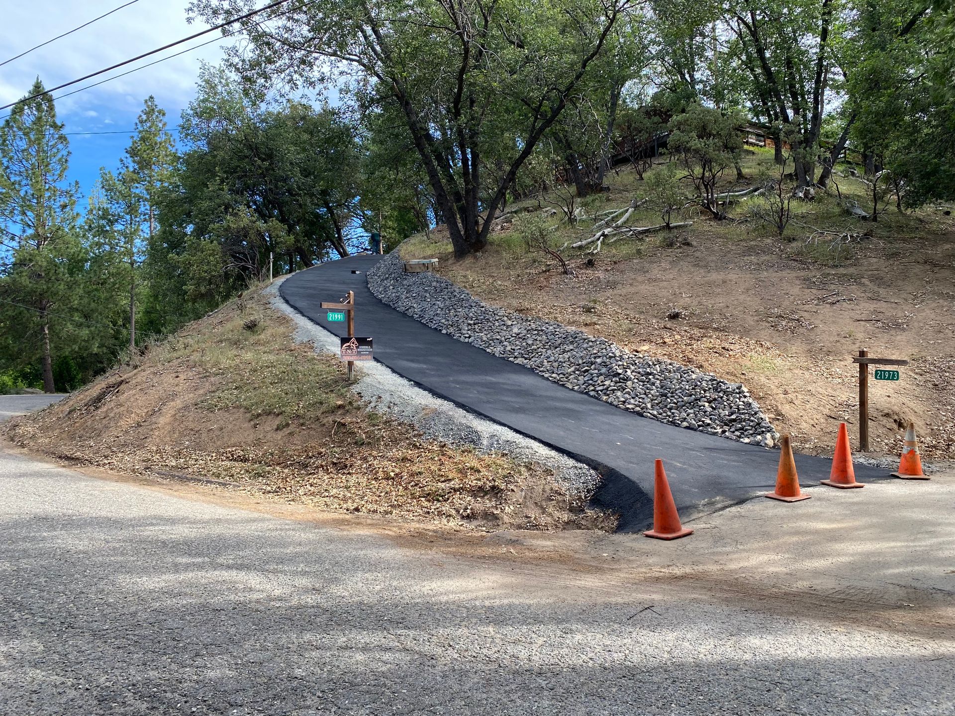 A paved, curving driveway slopes upward from a gravel road, lined with large rocks and orange traffic cones on the right.