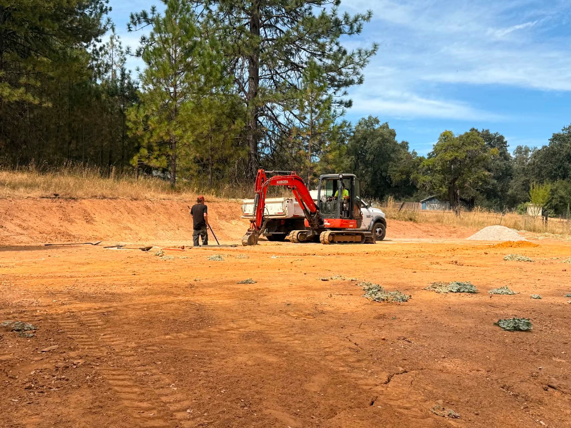 Excavator and worker on a dirt construction site with trees in the background