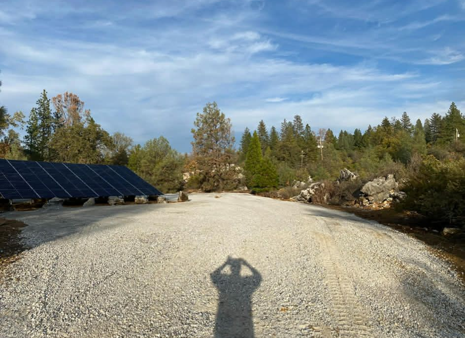 Ground-mounted solar panels sit on a gravel clearing beside a wooded area under a partly cloudy sky.