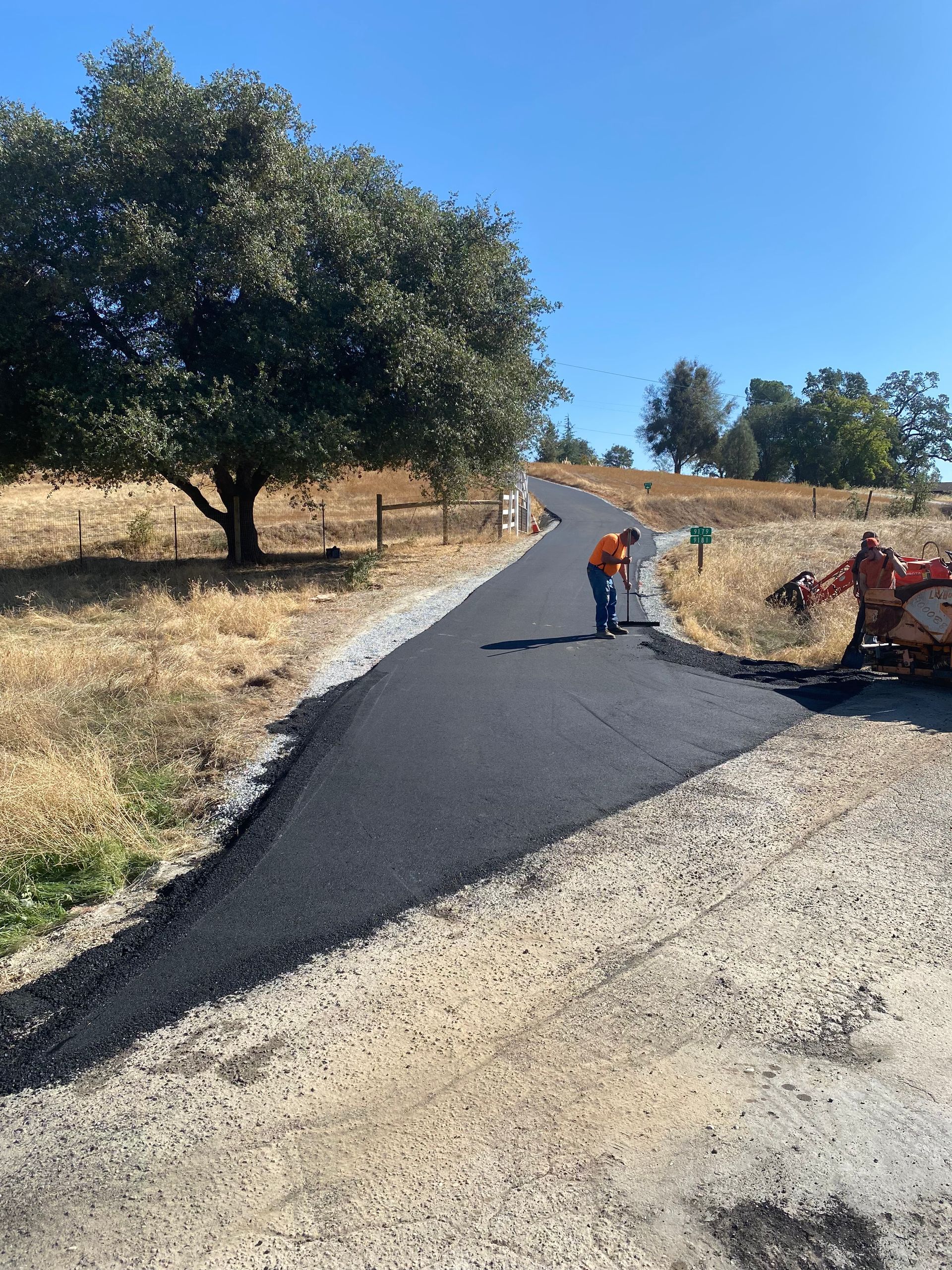 A worker in high-visibility orange clothing paves a new asphalt driveway leading uphill through a dry, rural field.