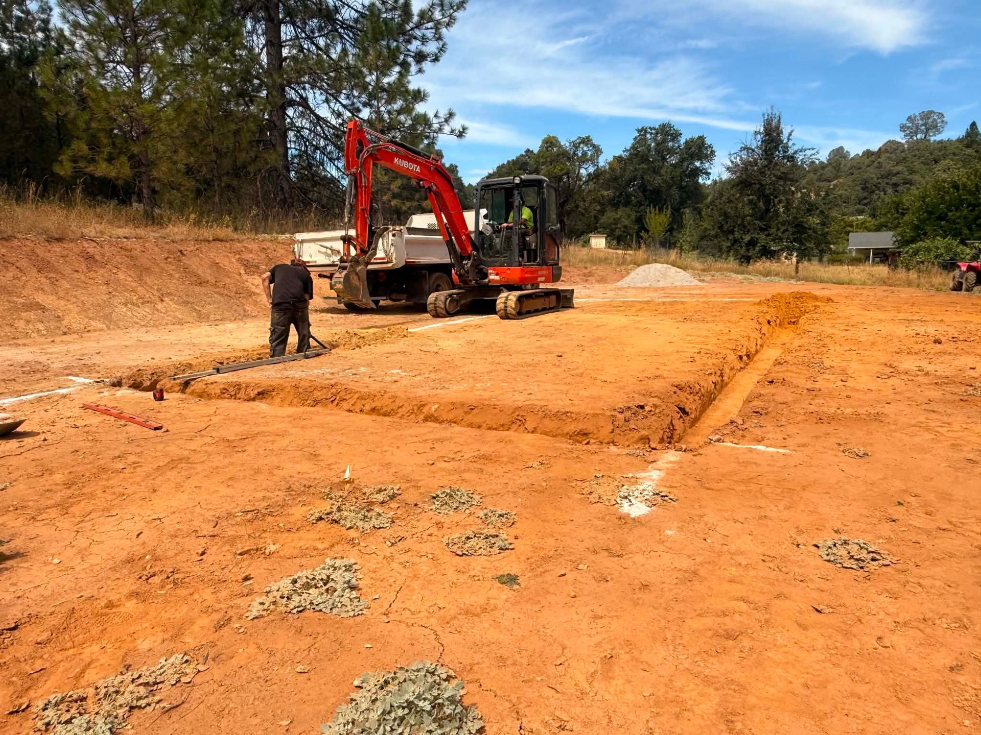 Excavator and worker grading a red dirt construction site under a blue sky