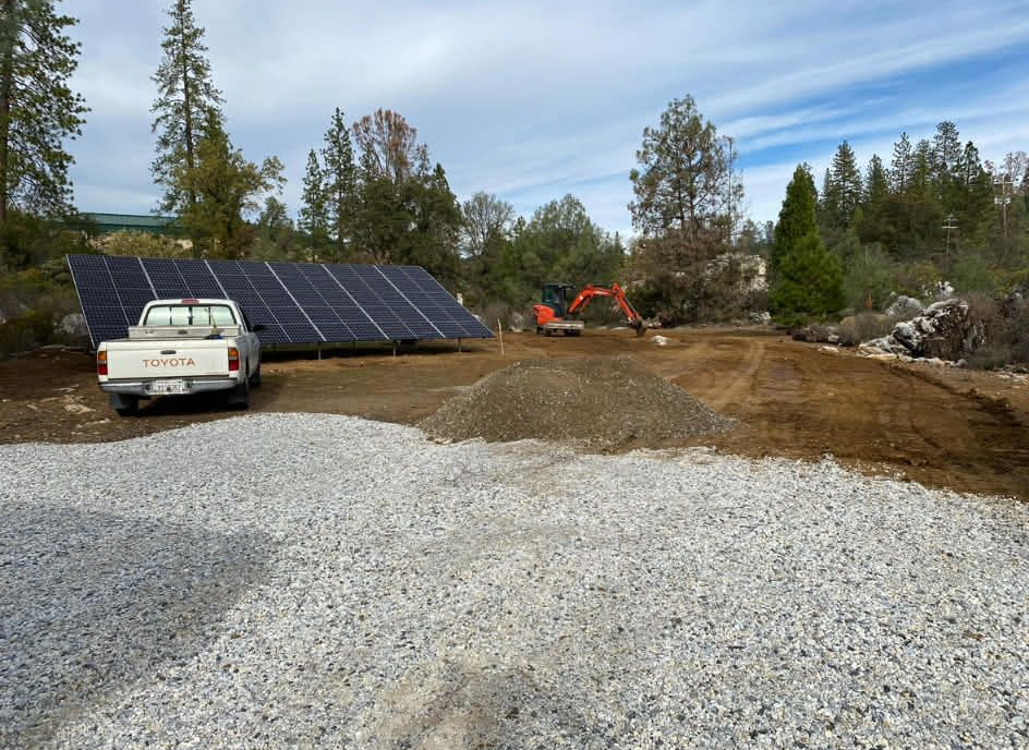 A white pickup truck parked in front of a ground-mounted solar array at a construction site with a small excavator.
