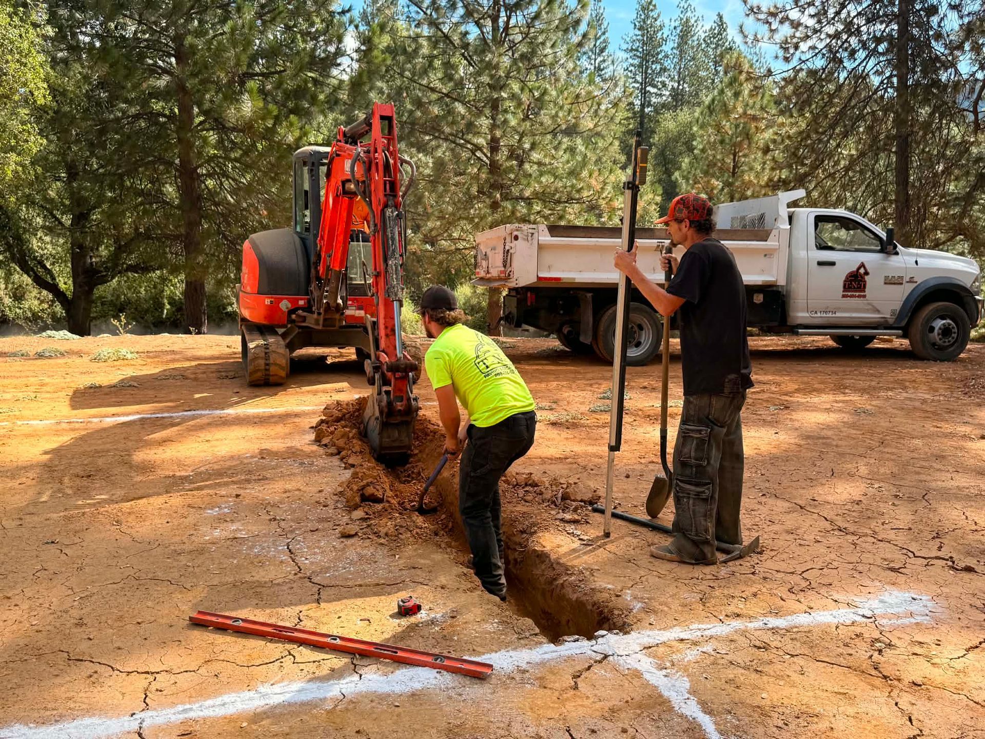 Workers dig a trench beside an orange excavator and white pickup in a forest clearing.