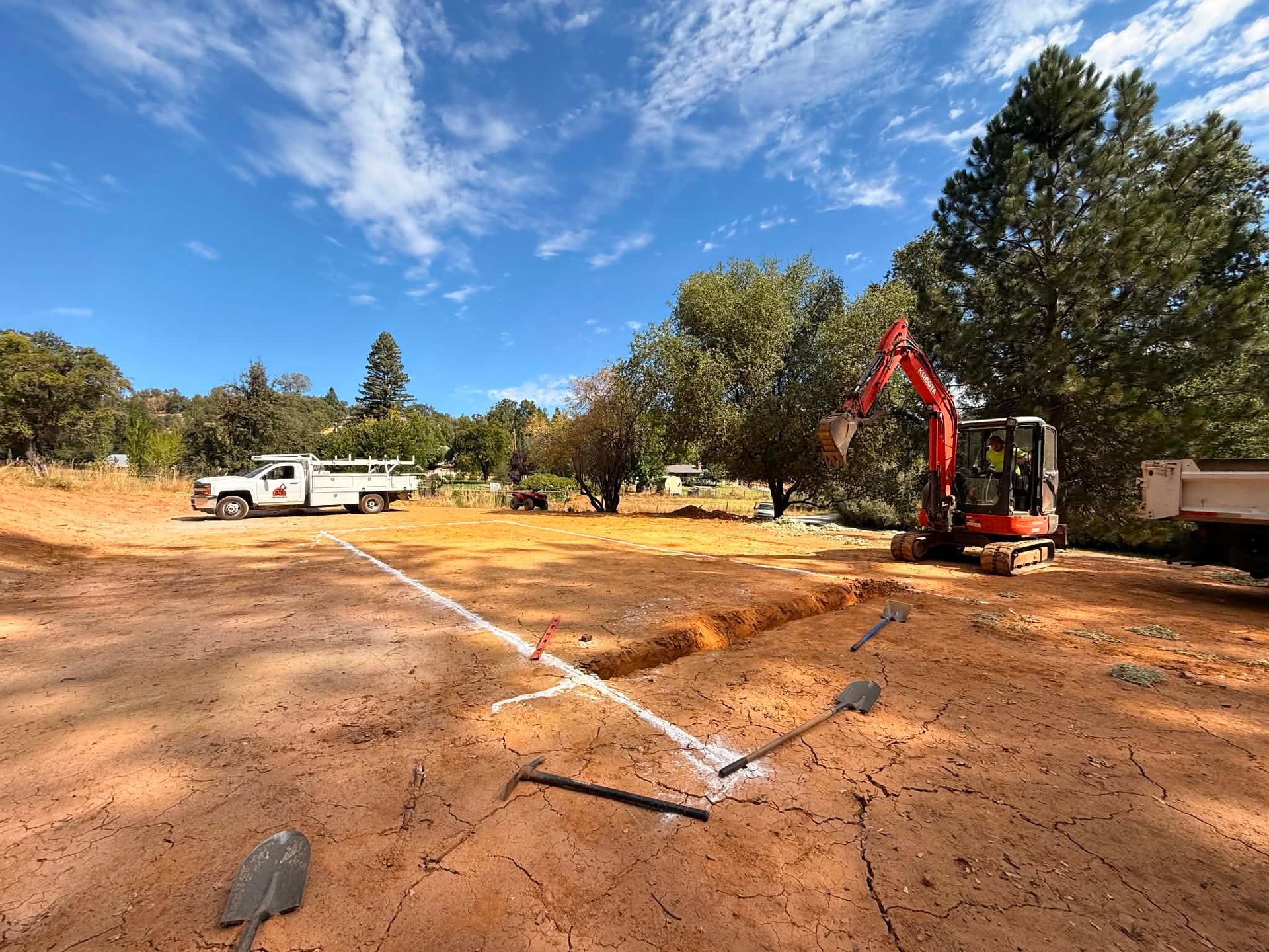 Excavator digging on a dusty lot with a white truck and trees under a blue sky