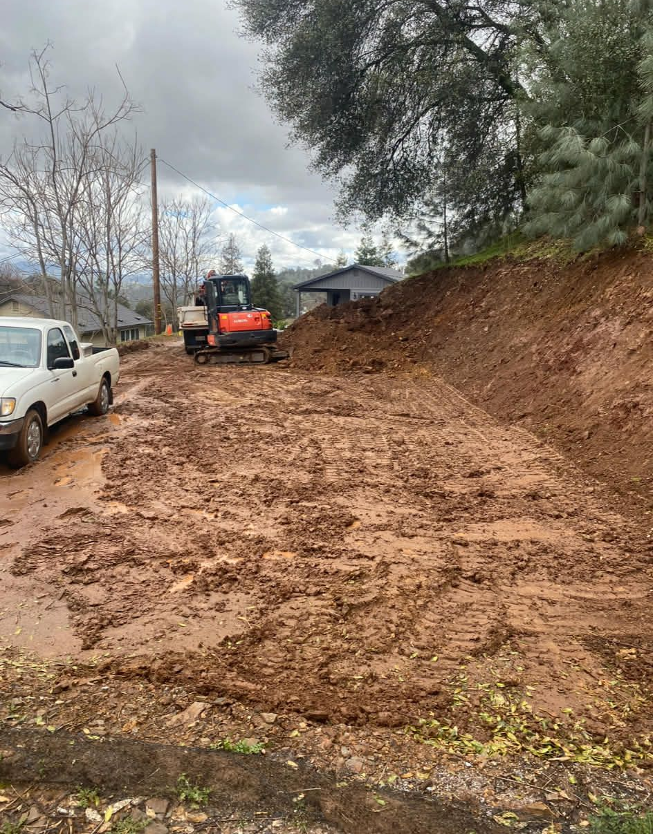 An orange mini-excavator sits on a muddy, excavated hillside near a white pickup truck on an overcast day.