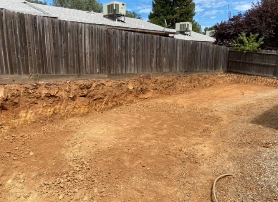 A backyard with a large, freshly excavated trench along a wooden fence, showing bare dirt and a leveled ground surface.