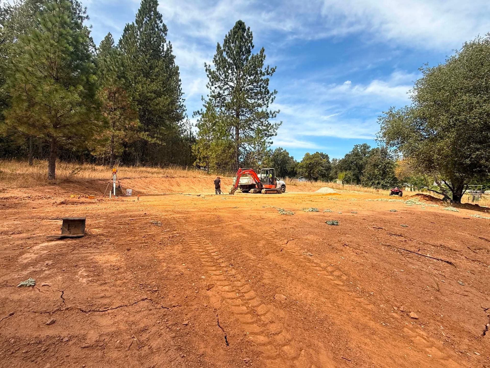 Dirt clearing with trees and a small red excavator under a blue sky