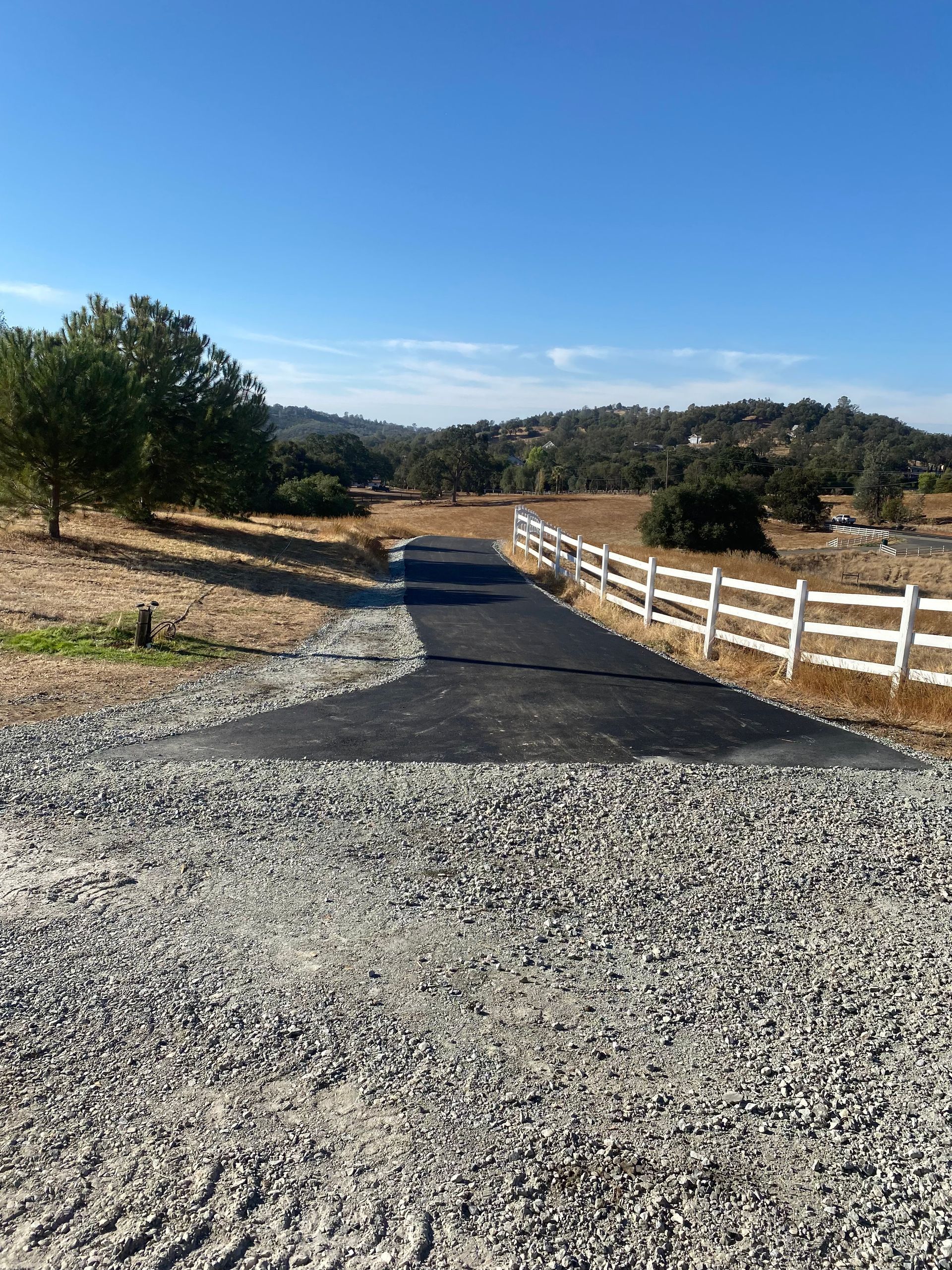 A paved driveway starts at a gravel area and leads into a field with trees and a white fence under a clear blue sky.