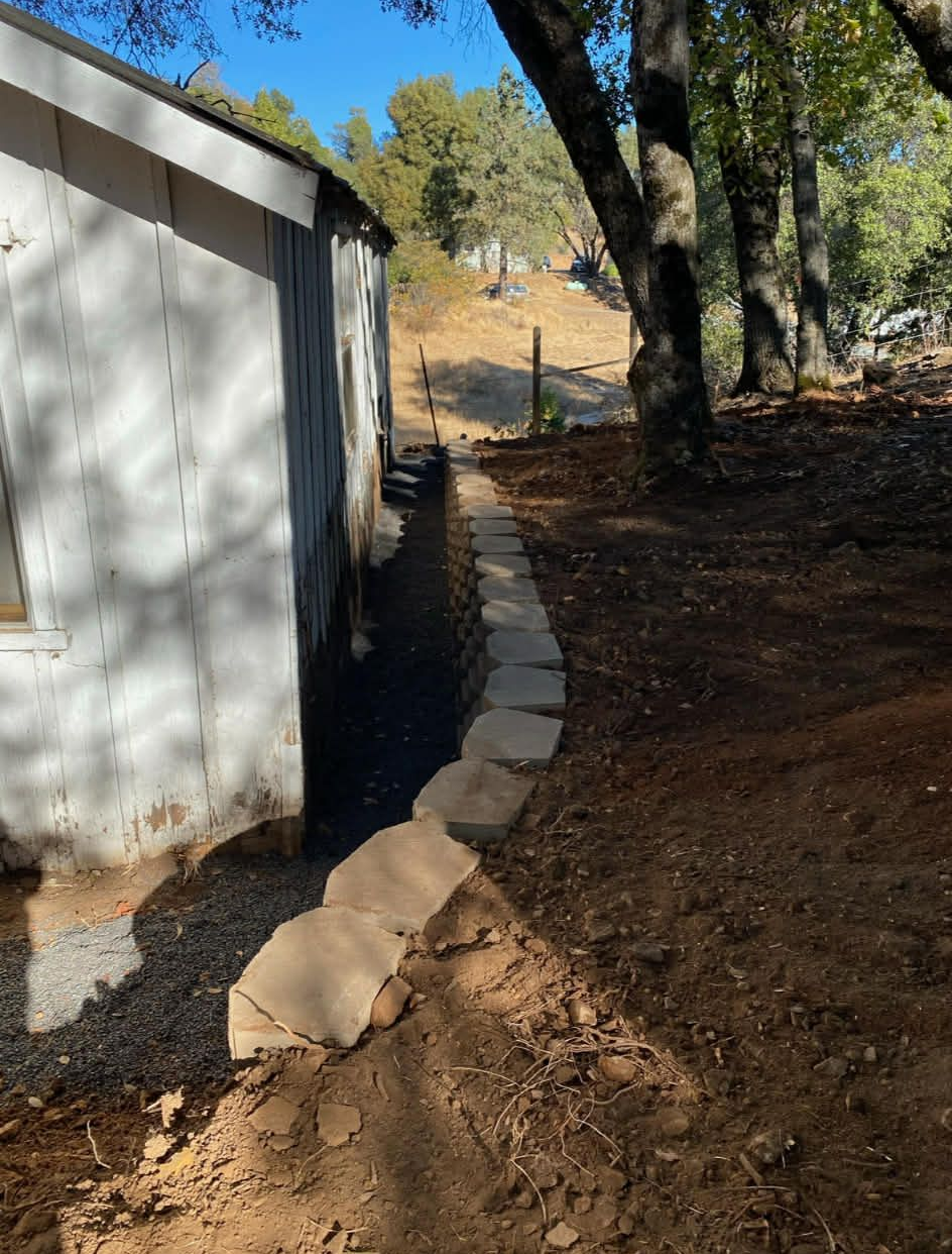 A white exterior building wall next to a stone border separating a gravel trench from a sloped dirt hillside.
