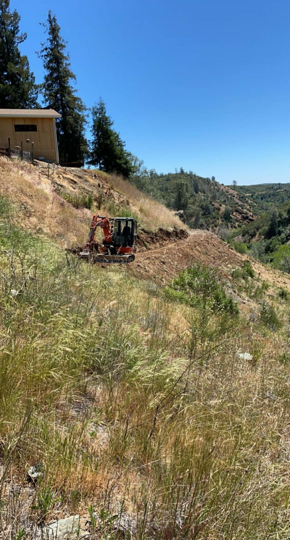 An orange excavator digs into a dry, grassy hillside near a building under a clear blue sky.