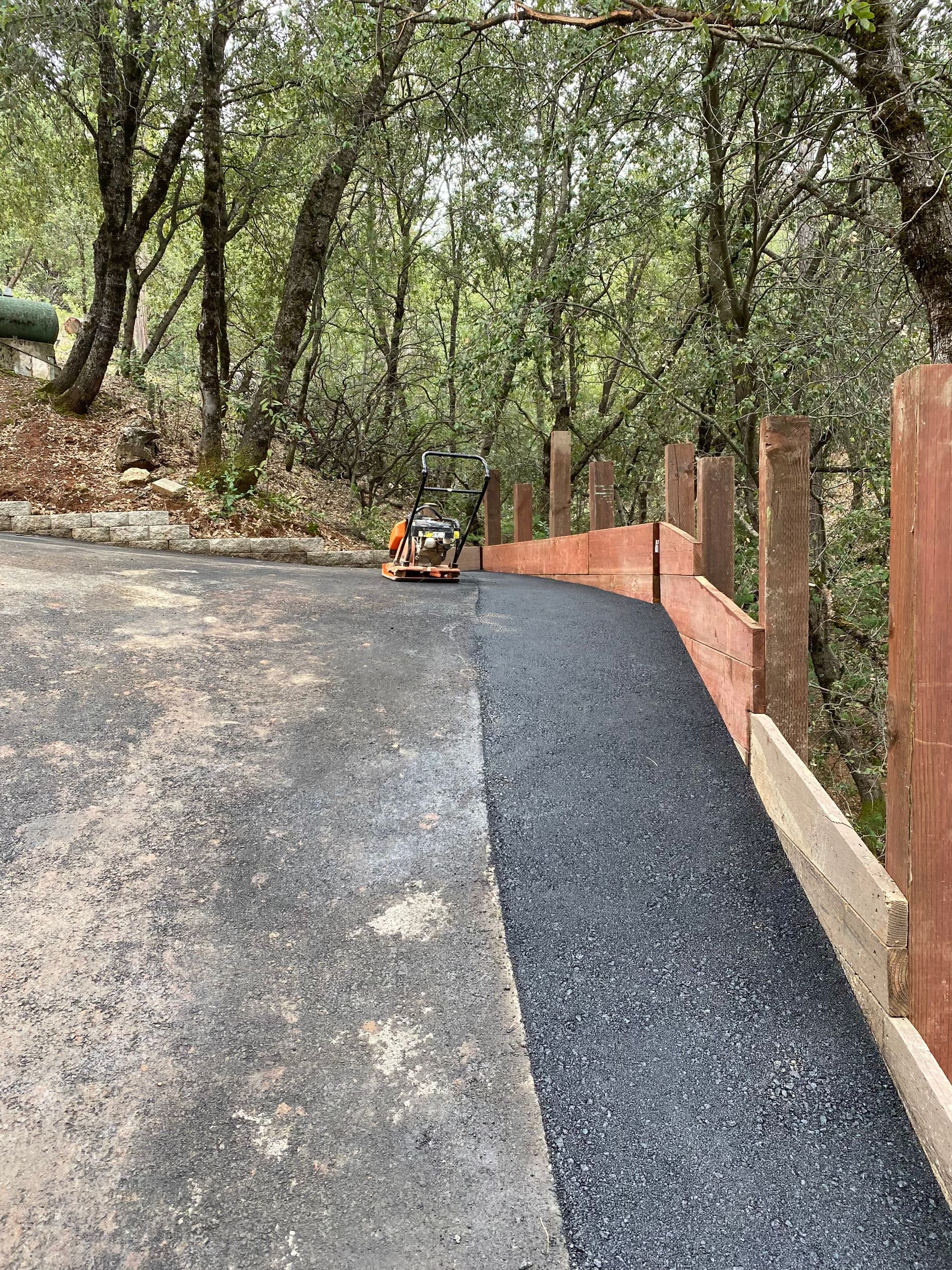 A path being paved with asphalt alongside a wooden retaining wall in a wooded area.