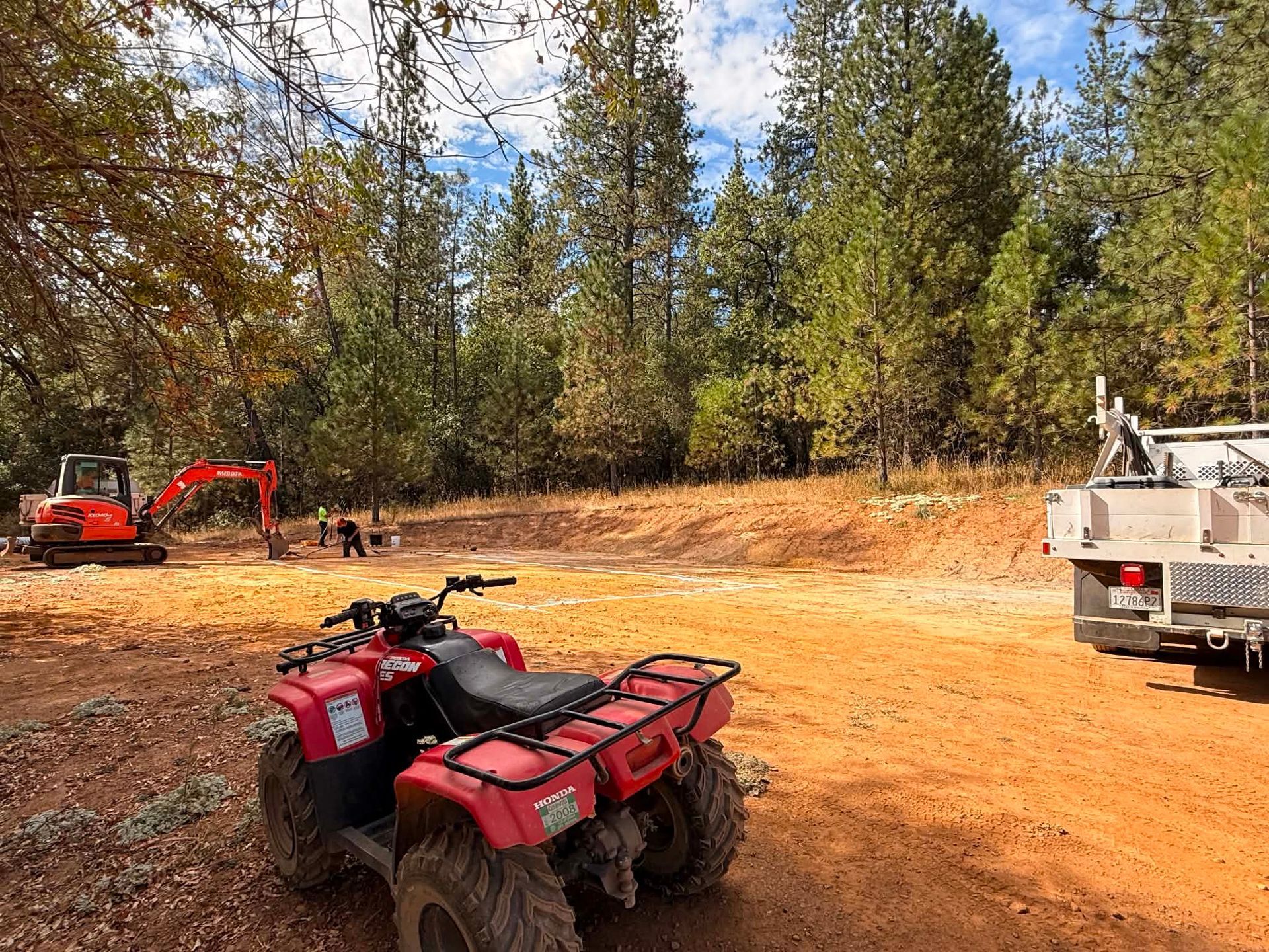 Red ATV parked on a dirt clearing with excavators and a dump truck in the background under trees.