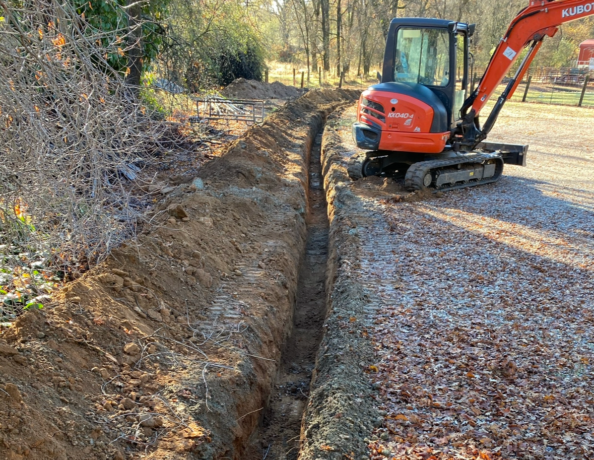 Orange Kubota excavator digging a long trench in a gravel driveway, alongside foliage and trees.