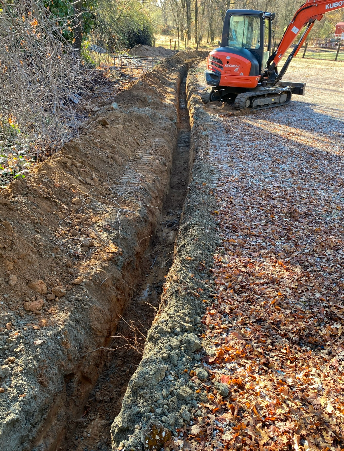 An orange mini excavator parked beside a long, freshly dug trench in a leaf-covered outdoor area.