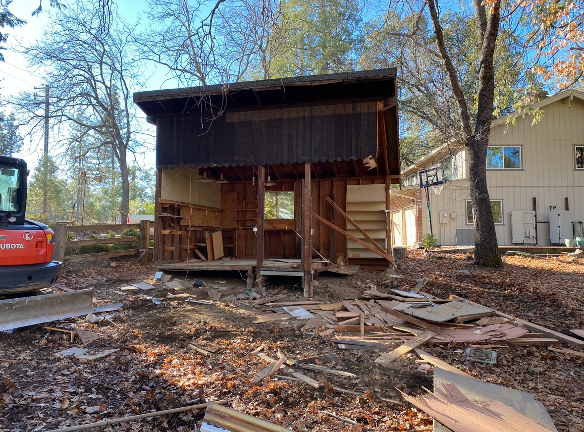 A small wooden building undergoing demolition, with debris on the ground and an excavator nearby in a wooded yard.