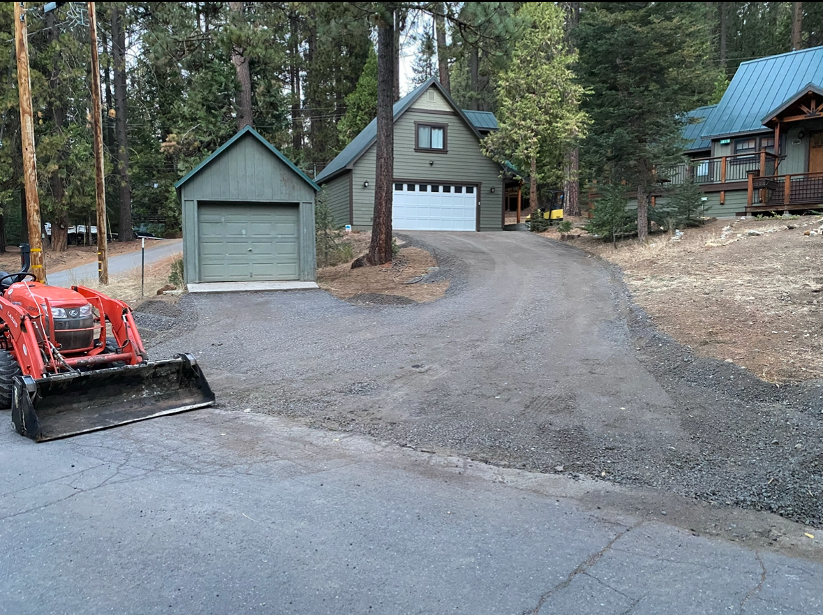 A red tractor with a front loader sits at the edge of a new gravel driveway leading to a two-story house and shed.