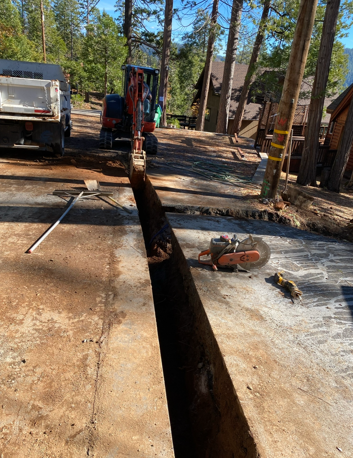 A construction site shows an orange excavator digging a long trench in a paved area, with a saw resting nearby.